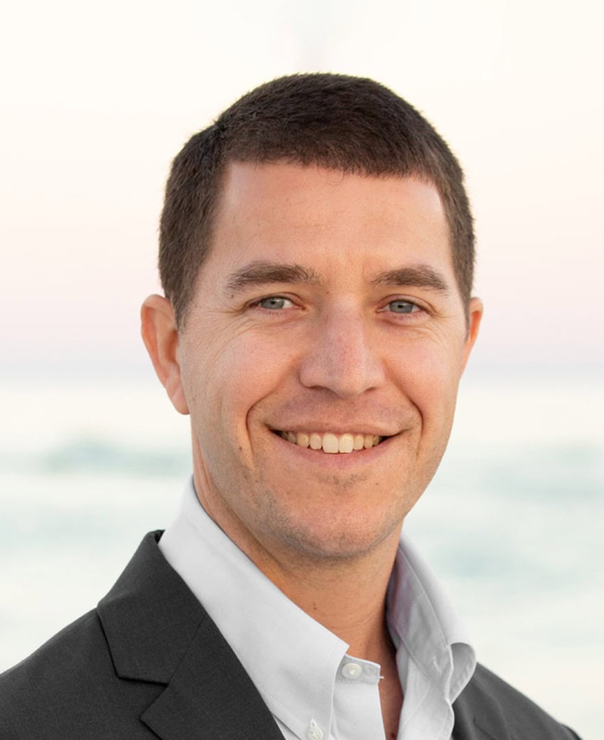 Dr. Carl Speer, board-certified podiatrist and creator of InvisiFoot™, smiling against a beach backdrop, emphasizing expertise in minimally invasive foot surgery.